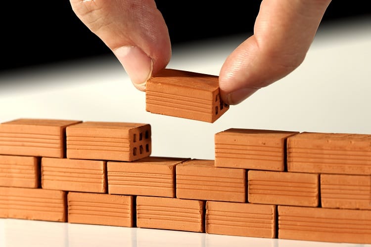 Large fingers building a tiny brick wall symbolizes using small investment ideas to cultivate wealth.